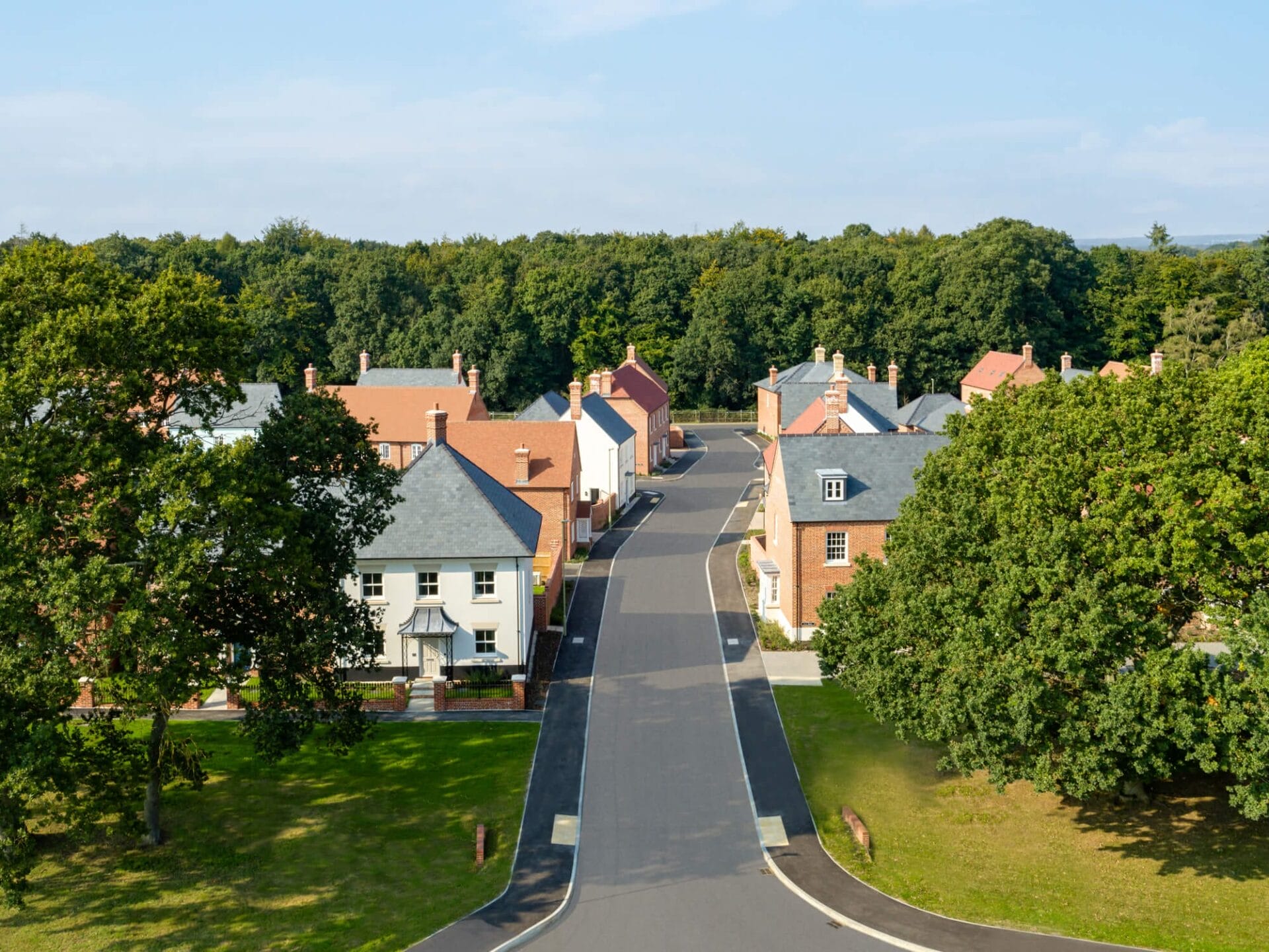 A scenic aerial view of a residential development featuring traditional-style homes with red brick and white facades, slate and tiled roofs, and chimneys. The neighborhood is surrounded by lush green trees, creating a peaceful and picturesque setting. A smoothly paved road winds through the community, lined with neatly maintained lawns and mature trees, enhancing the suburban charm.