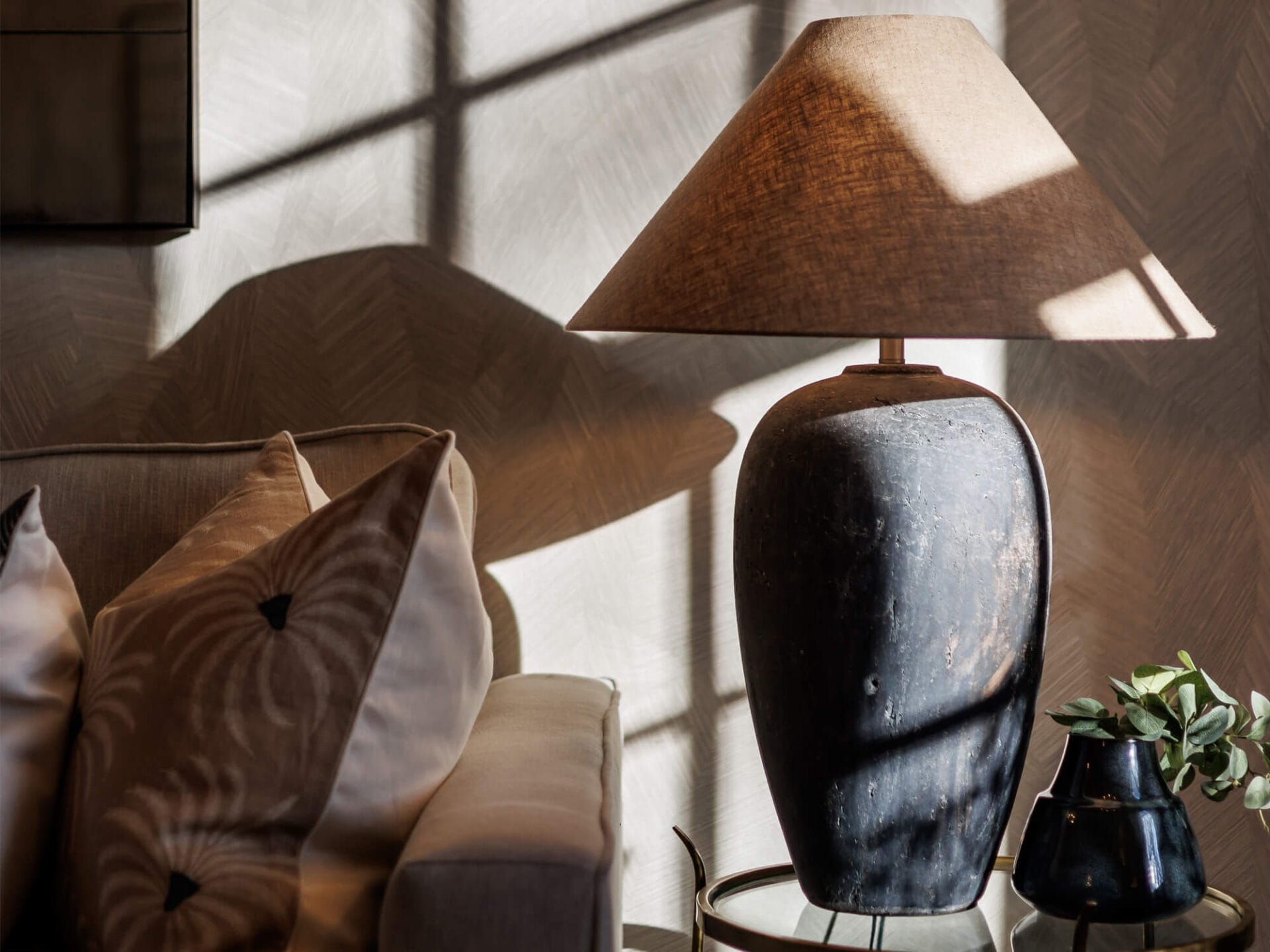 A cosy interior setting featuring a rustic table lamp with a textured, dark ceramic base and a beige lampshade, casting geometric shadows on the wall. The lamp sits on a round glass-top side table beside a dark vase with green foliage. A plush beige sofa with decorative pillows, including one with a radial embroidered pattern, is positioned nearby, adding to the warm and inviting atmosphere.