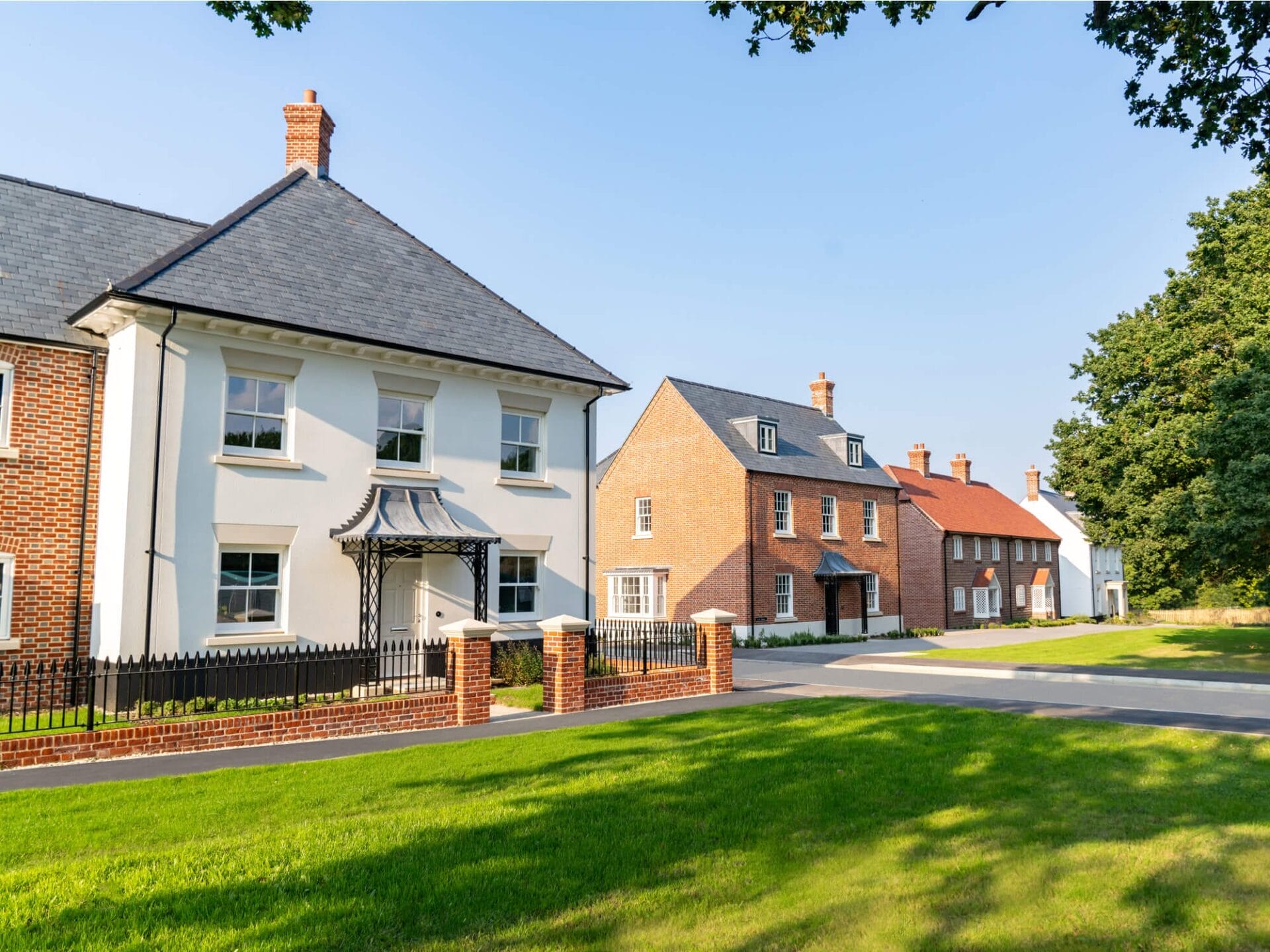 A row of elegantly designed traditional-style homes in a peaceful residential neighborhood. The foreground features a white house with a slate roof, decorative iron porch canopy, and a black wrought iron fence with brick pillars. Adjacent homes showcase red brick and white facades, pitched roofs, and chimneys, exuding a timeless architectural charm. Lush green lawns and mature trees frame the setting under a bright blue sky.